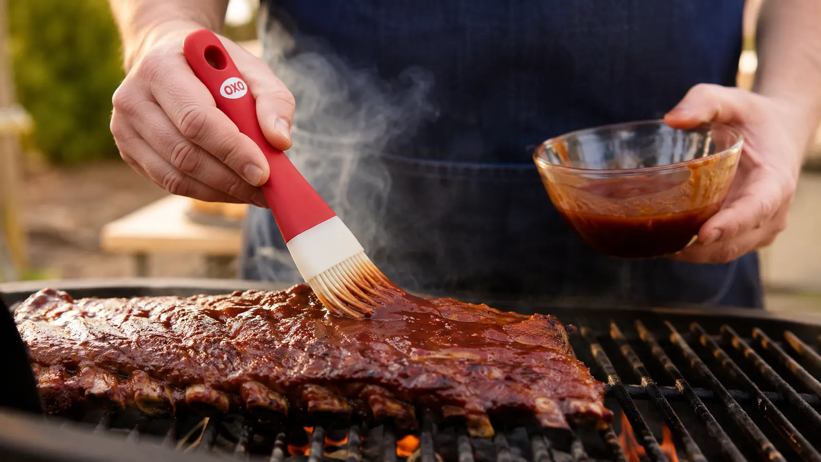 Hand holding the OXO Good Grips silicone basting brush and painting BBQ sauce across a rack of ribs on the grill