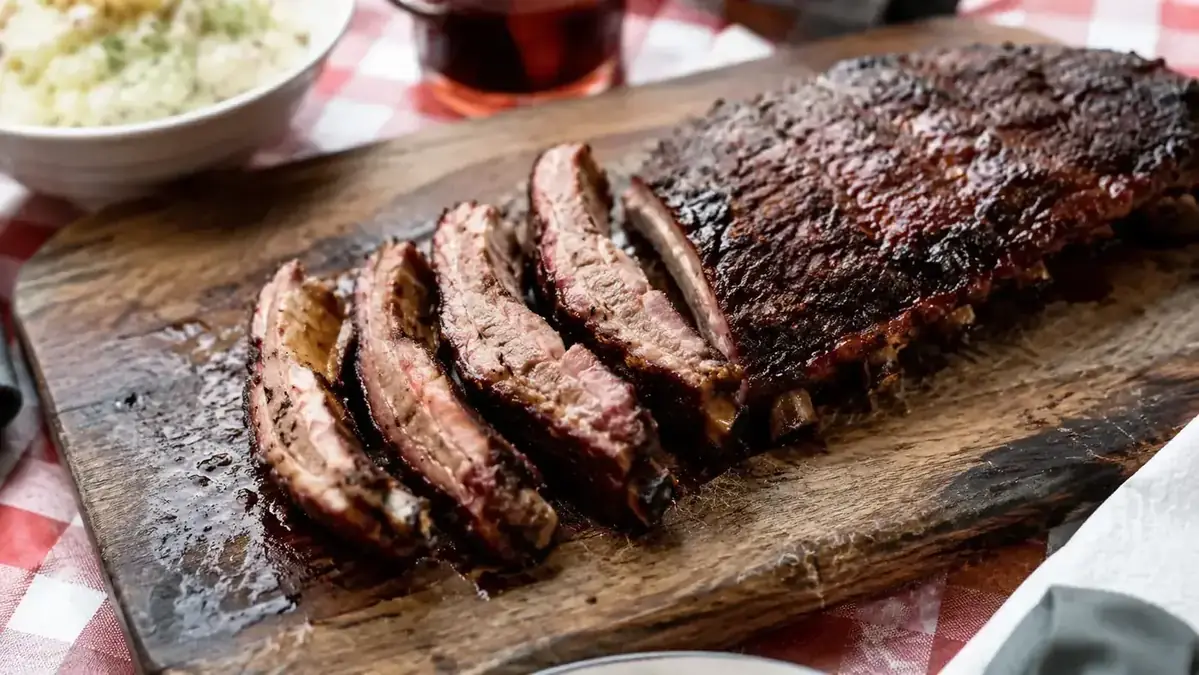 Finished rack of glazed ribs sliced and resting on a wooden cutting board, dark mahogany crust, backyard table setting