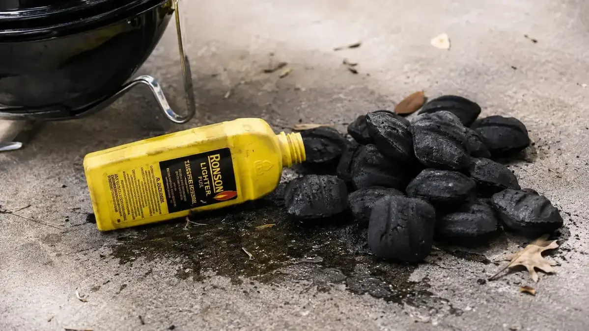 Bottle of lighter fluid next to a charcoal pile on a grill, cautionary visual contrasting with the clean chimney starter method