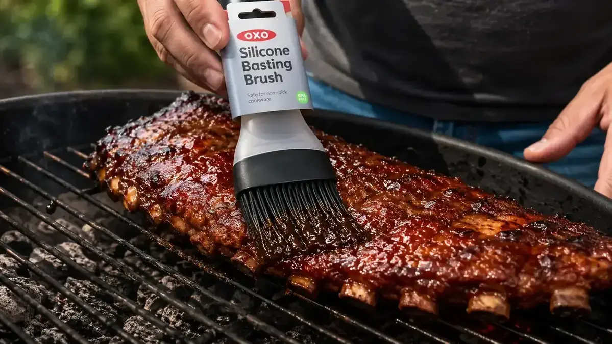 Close-up of OXO silicone basting brush bristles pressing into the surface of a rack of ribs on a hot grill grate