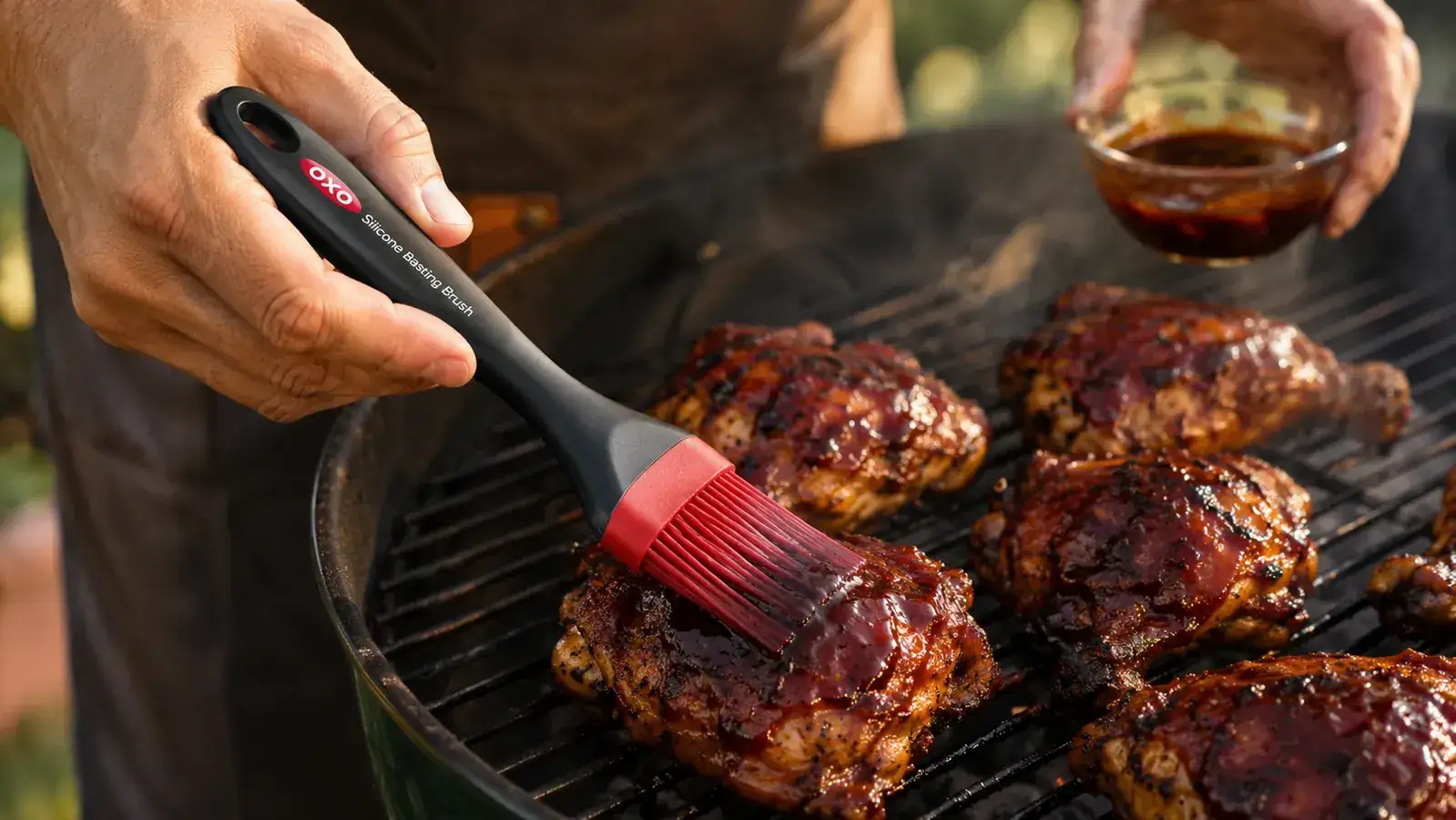 Hand using a red silicone basting brush to apply BBQ sauce to chicken thighs on a gas grill grate