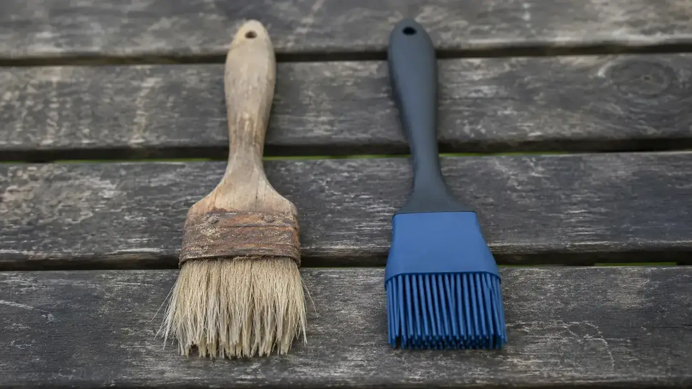 Old frayed bristle pastry brush next to a modern red silicone basting brush on a wooden table