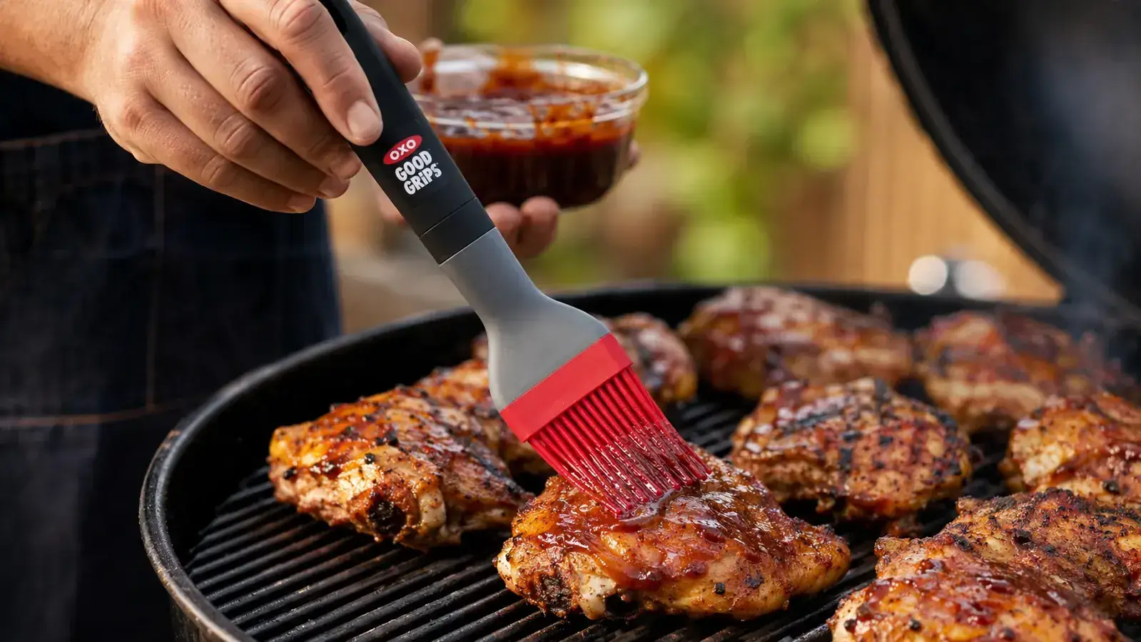 Hand using the OXO Good Grips silicone basting brush to apply BBQ sauce to chicken thighs on a grill