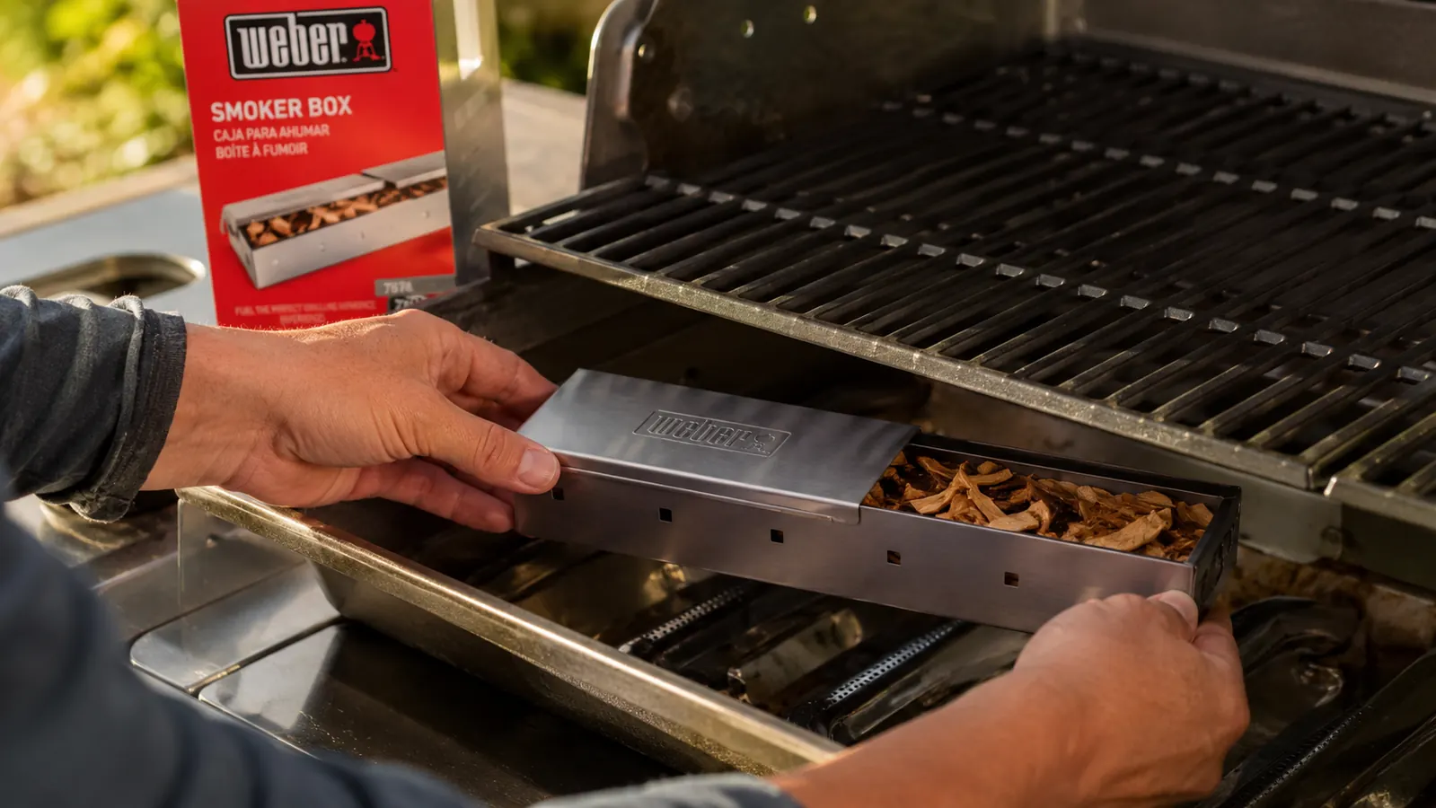 Hand placing Weber smoker box filled with hickory wood chips over a gas burner on a stainless gas grill